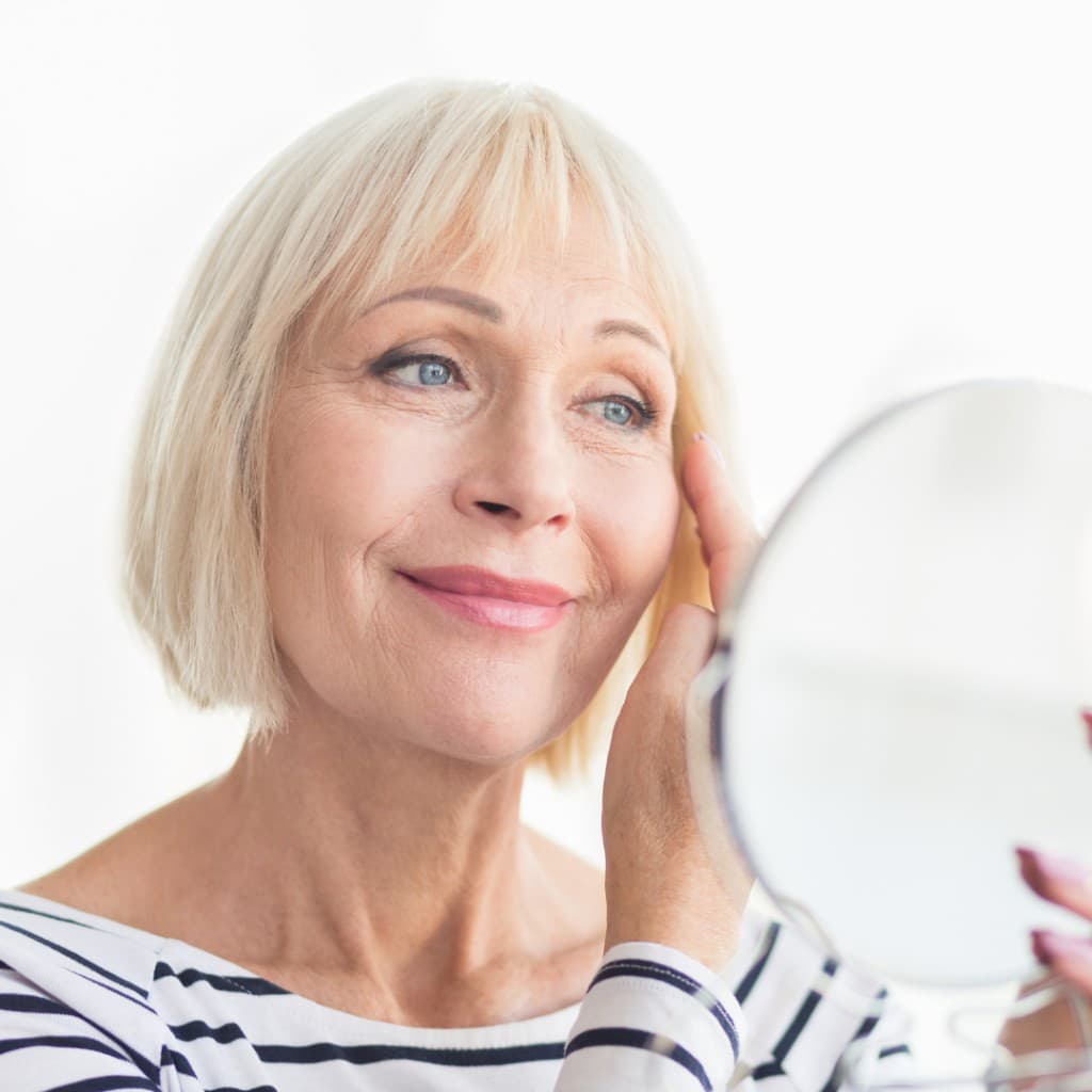 Mature woman using a small mirror while gently touching the outer eye area